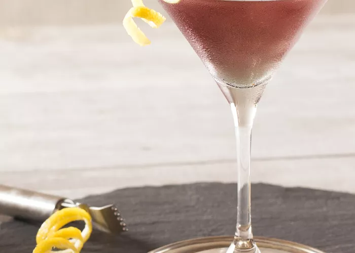 A glass of Purple Haze Martini, garnished with a  twisted lemon peel on the rim of the glass, is placed  on a steel coaster. Alongside it, there is another t wisted lemon peel and a stainless steel Lemon  Zester Grater with a Channel Knife and Hanging  Loop on a round wooden coaster.