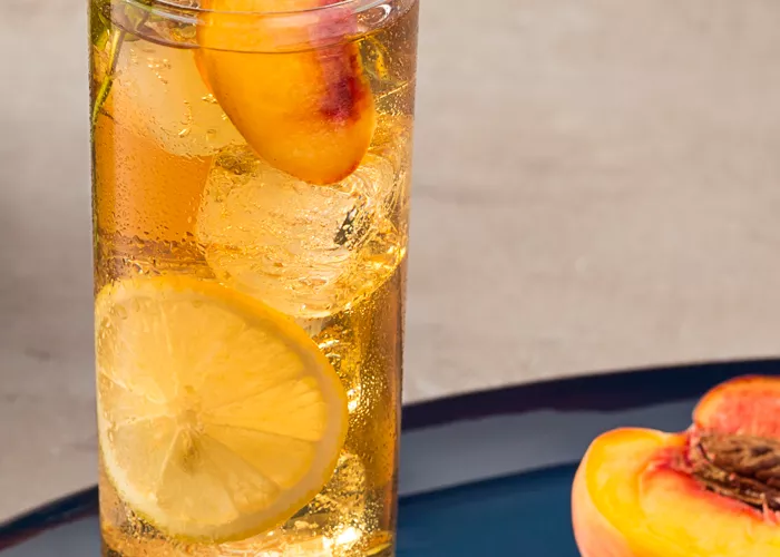 A highball glass of Peach Iced Tea, garnished with lemon wheel, peach slices and mint leaves, accompained by halved peach.
