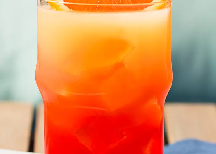 A glass of Tequila Sunrise, garnished with an orange wedge and a straws, is placed on a blue cloth