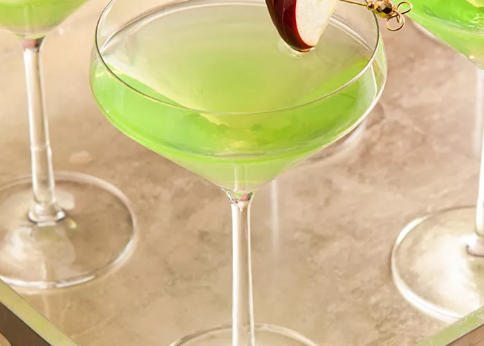 Four glasses of Appletini, each garnished with an  apple slice pierced on a cocktail pick on the rim  of the glass, are arranged in a wooden tray.