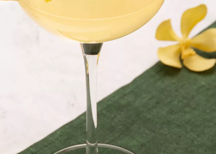 A glass of Bee's Knees garnished with lemon peelon a green cloth alog with the yellow flowers  and bowl of lemons 