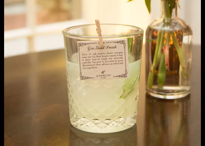 A glass of Gin Basil Smash is garnished with basil leaves and ice cubes, with a paper note attached to the rim of the glass with a clip, alongside a jar filled with water containing a basil plant.