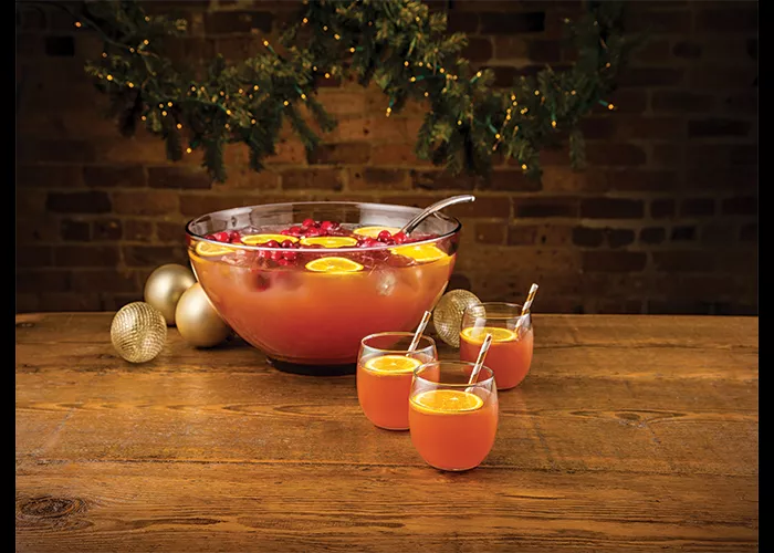 Three glasses of Holly Jolly Punch garnished with orange slice and straws alongside pot of Holly Jolly Punch 