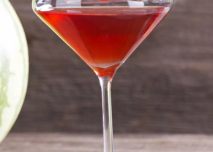 A glass of Melon Creek Martini, sitting on a cloth, is garnished with spiked watermelon balls pierced on steel cocktail picks, placed on the glass rim