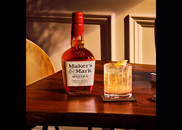 Glass of Maker's Mark® Gold Rush on a coaster alongside bottle of Maker's Mark Whiskey