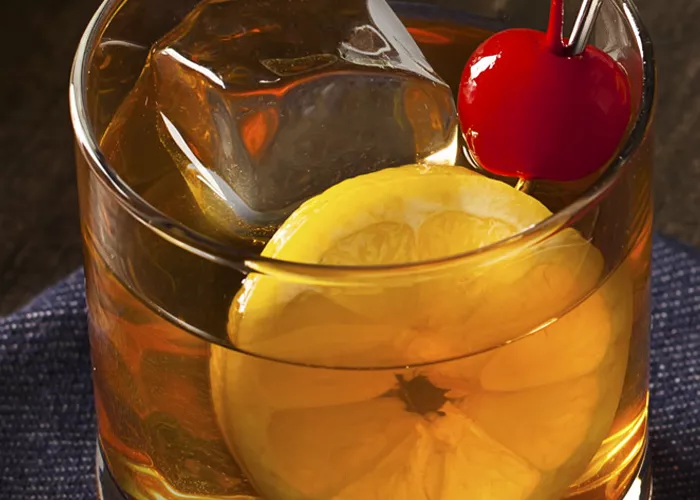 Glass of Maker's Mark® Sour garnished with ice, lemon slice and cherry placed on a blue cloth.