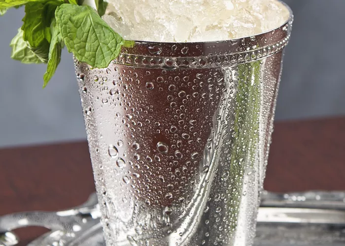 Silver glass of Maker's Mark Mint Julep with straws and crushed ice, garnished with mint leaves, alongside some additional mint leaves on a silver tray.