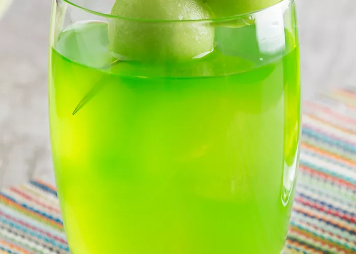 A glass of bright green drink garnished with two melon balls skewered on a toothpick, placed on a colorful striped mat.