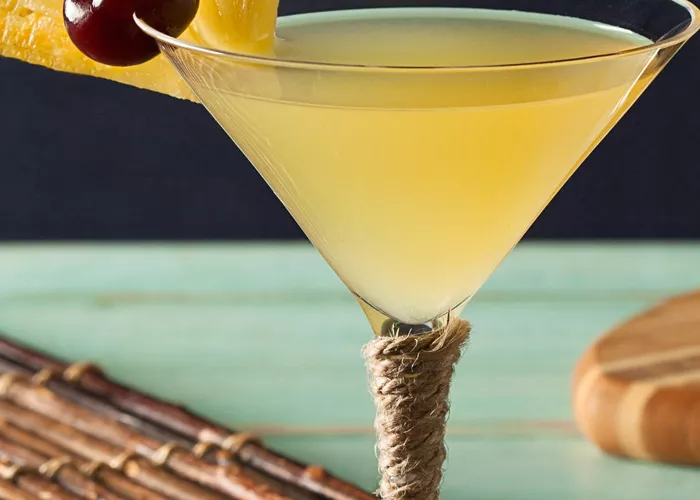 A glass of Pina Colada Martini garnished with a slice of pineapple and a cherry on the rim, placed on a wooden mat