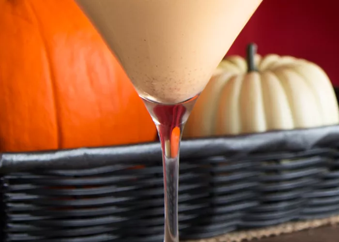A glasses of Pumpkin Pie Martini garnished with whipped cream and Pumpkin Spice, placed on a small piece of cloth. Nearby, a tray of pumpkin on a cloth