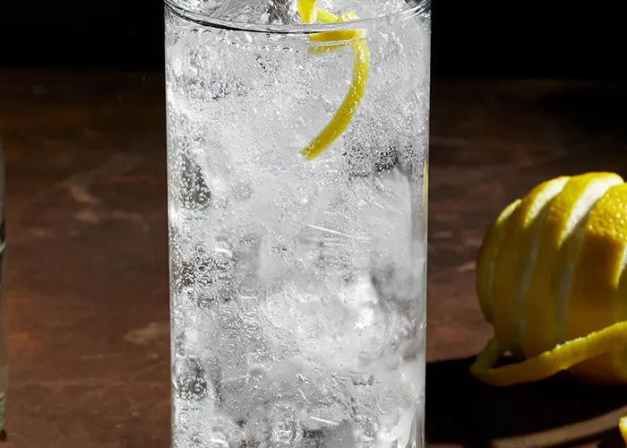 A glass of Roku® Gin Sonic with ice, half-filled water bottle, half-filled green glass, artisticallly chopped lemon, and copper spoon on the table.