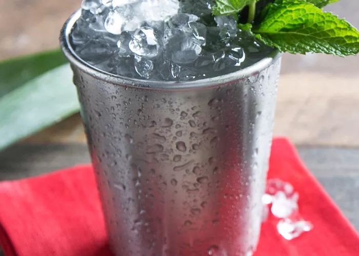 A steel glass of Mexican julep, garnished with crushed ice and mint leaves, sits on a red cloth with an ice cube placed on top, all positioned on a table.