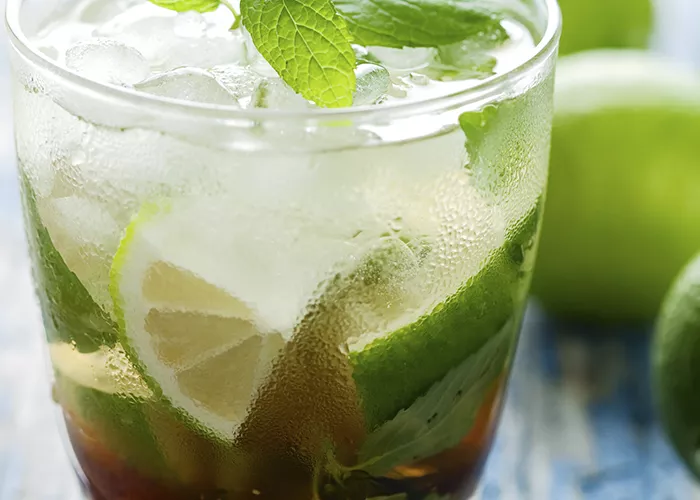 A glass of Banana Cinnamon Mojito garnished with fresh spanked mint and some lemon slices. alongside few lemons, some mint leaves and a steel spoon filled with sugar.