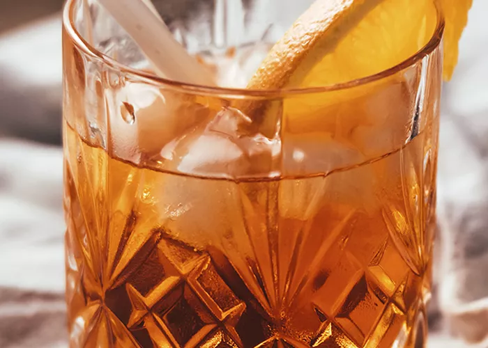 A glass of Coconut Old Fashioned with coconut water ice cubes, garnished with an orange slice on the rim, and two straws