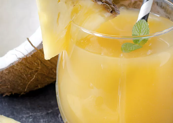 A glass of Delusion garnished with a piece of pineapple and few mint leaves along with a straw alongside a halved coconut and  a pineapple.