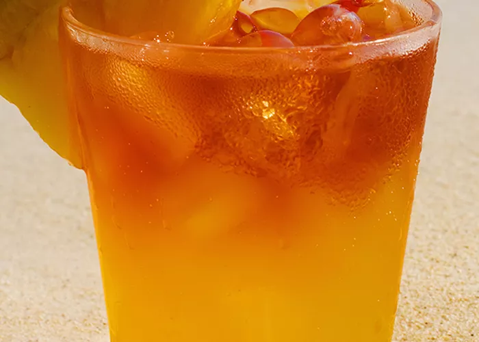 A glass of Mango Hurricane, garnished with an orange wedge and a cherry, along with a pineapple slice and a cocktail umbrella on the rim of the glass with a straw.