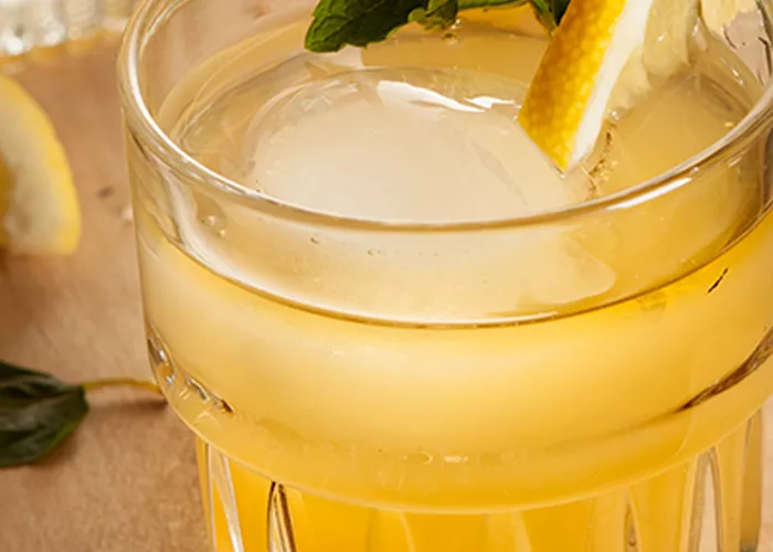 A glass of Nil Fashioned garnished with a slice of lemon and mint leaves, some of which are fallen on the wooden table.