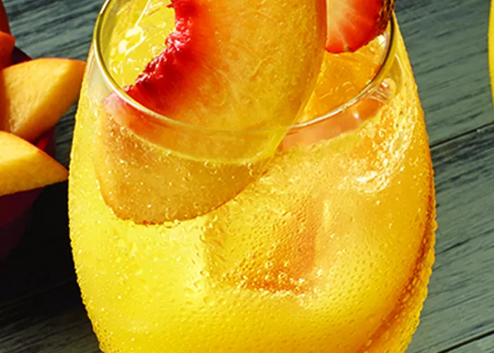 A glass of Peach on the Beach garnished with a peach slices and half cut strawsberry with staw. alongside a bowl of sliced peaches.