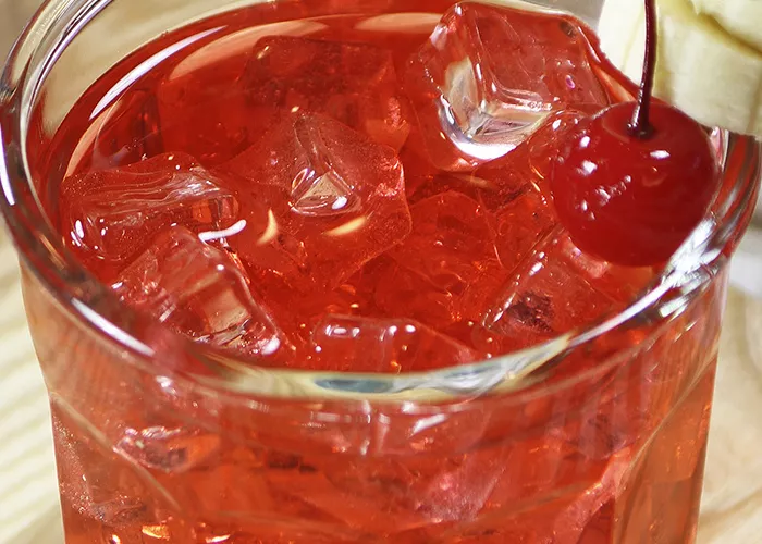 A glass of DeKuyper® Banana Split, garnished with ice cubes, a cherry, and banana slices pierced on a toothpick, is placed on the rim of the glass