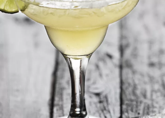 A glass of Elderflower Margarita garnished with ice cubes and a wheel of lemon on rim of glass