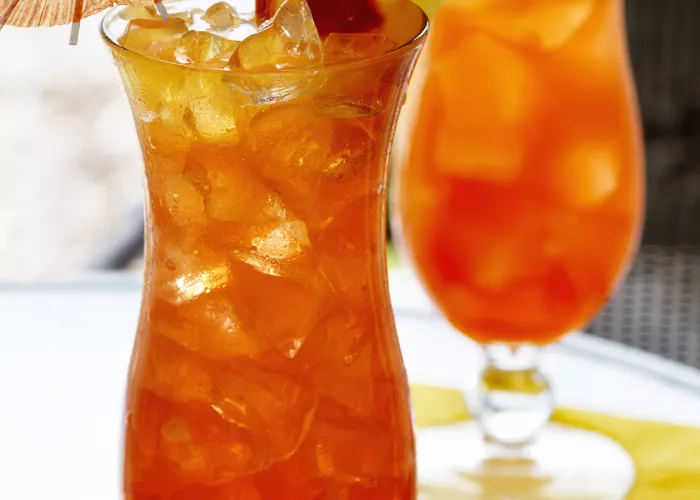 Two glass of Mai Time, garnished with ice cubes, a cherry, orange and  pineapple slice on the rim of the glass, and a cocktail umbrella