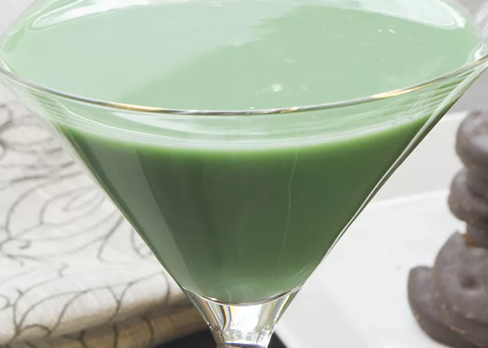 "A glass of Mintini Martini is placed alongside a white  cloth, while on the other side, there are a few  cookies arranged on a white tray."