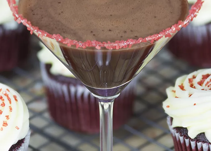 A glass of Red Velvet Cake Martini garnished with sugar mixture on rim of glass along side few cupcakes