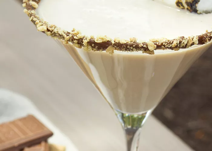 Glass of Smore-Tini garnished with a toasted marshmallow and rimmed with crushed graham crackers and melted chocolate, alongside some chocolate and marshmallows.