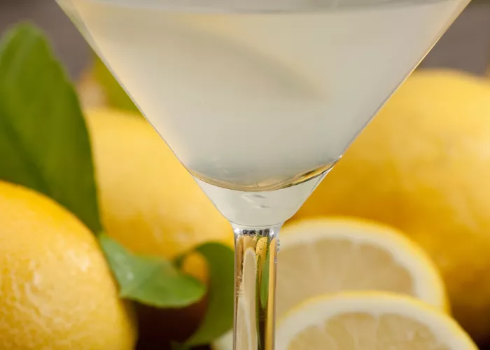 A martini glass of Kiwi Zest Martini, garnished with a lemon wedge and rimmed sugar. Alongside couple of lemons, lemon slices, and leaves.