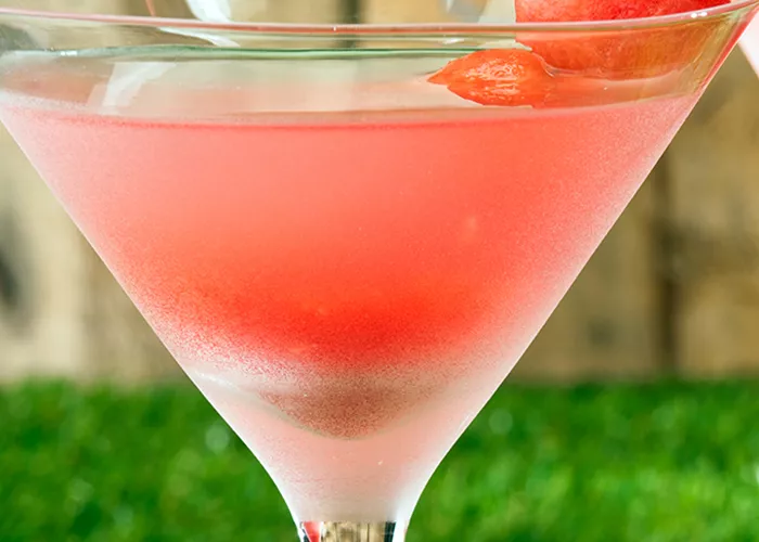 A martini glass of Pinnacle® Summer Breeze garnished with a watermelon wedge on cocktail pick placed on a green grass surface.