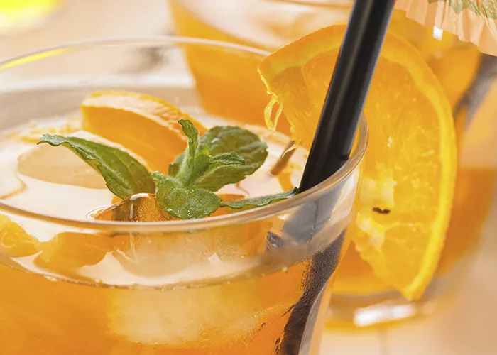 Two glasses of Sunset Tea garnished with an orange slice or apple spear dipped in cinnamon and mint leaves along with a straws and cocktail umbrella.