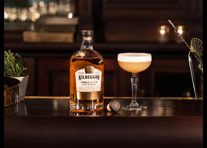 A bottle of Kilbeggan® Brosna Sour, accompanied by a glass of Kilbeggan® Brosna Sour, is placed on a black table.