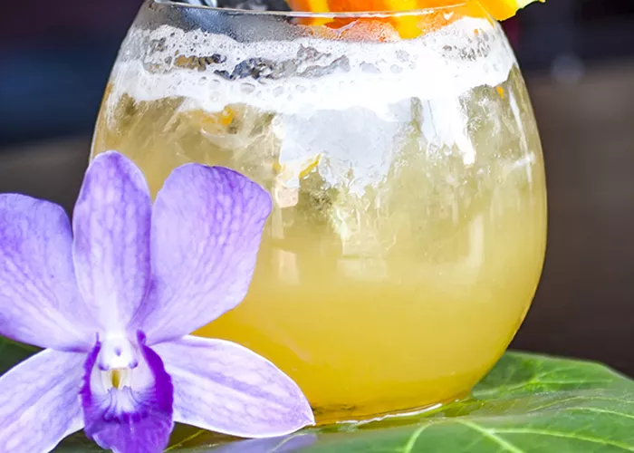 A glass of Mango Rum Margarita garnished with orange slice and two strawss with a purple flower on a leaf