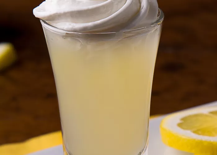 A glass of Ooh La La Lemon topped with whipped cream sits on a coaster, accompanied by lemon slices
