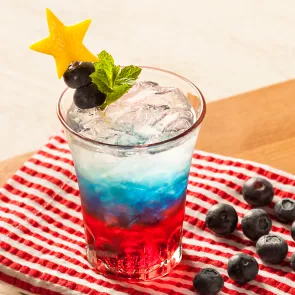 Glass of Citrus-Spangled Spritzer garnished with mint leaves, lemon rind star and berries alongside some blueberries  on red white striped cloth