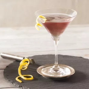A glass of Purple Haze Martini, garnished with a  twisted lemon peel on the rim of the glass, is placed  on a steel coaster. Alongside it, there is another t wisted lemon peel and a stainless steel Lemon  Zester Grater with a Channel Knife and Hanging  Loop on a round wooden coaster.