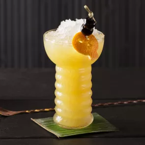 Glass of Zombie garnished with crushed ice, orange peel and cherries on coaster alongside a bar spoon