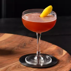 A glass of Black Sunset garnished with lemon peel on coaster on a round wooden tray