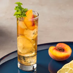 A highball glass of Peach Iced Tea, garnished with lemon wheel, peach slices and mint leaves, accompained by halved peach.