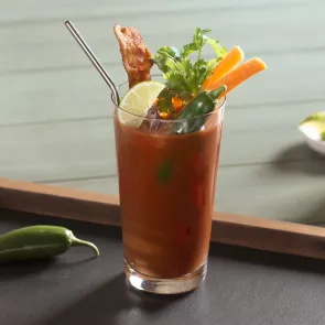 Glass of Sunday Funday Bloody Mary garnished with lime wedge, cilantro, becon slices on wooden tray alongside glass with becon slices