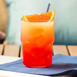 A glass of Tequila Sunrise, garnished with an orange wedge and a straws, is placed on a blue cloth