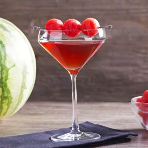 A glass of Melon Creek Martini, sitting on a cloth, is garnished with spiked watermelon balls pierced on steel cocktail picks, placed on the glass rim