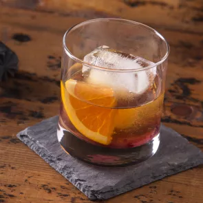 A glass of Peach Old Fashioned garnished with orange wedge on rock coaster