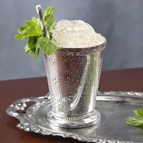 Silver glass of Maker's Mark Mint Julep with straws and crushed ice, garnished with mint leaves, alongside some additional mint leaves on a silver tray.