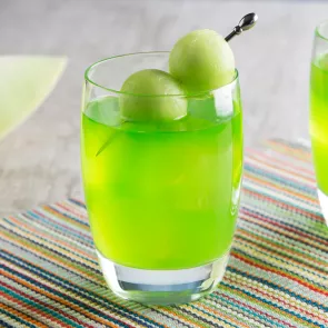 A glass of bright green drink garnished with two melon balls skewered on a toothpick, placed on a colorful striped mat.