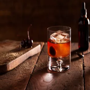 A glass of Classic Old Fashioned, garnished with a cherry and an ice cube, sits alongside cherries on a wooden plank placed on a wooden table.