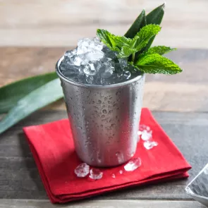 A steel glass of Mexican julep, garnished with crushed ice and mint leaves, sits on a red cloth with an ice cube placed on top, all positioned on a table.