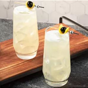 Two glasses of Tokyo Collins garnished with lemon peel and black cherries on a wooden try on the kitchen slab