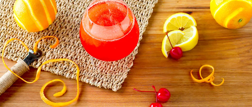 A glass of Sunset Daiquiri placed on jute mat, Along with couple of lemon slices, peeled orange, and cherries. A peeler  wit peeled orange and few half peeled oranges nearby. 