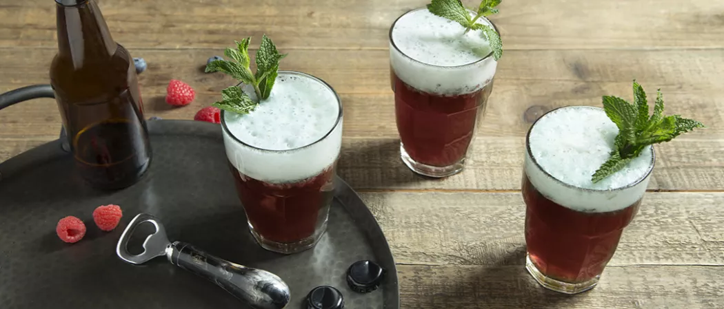 Three glasses of Berry Good Shandy garnished with mint leaves one on tray  alongside some bottle caps and a bottle opener